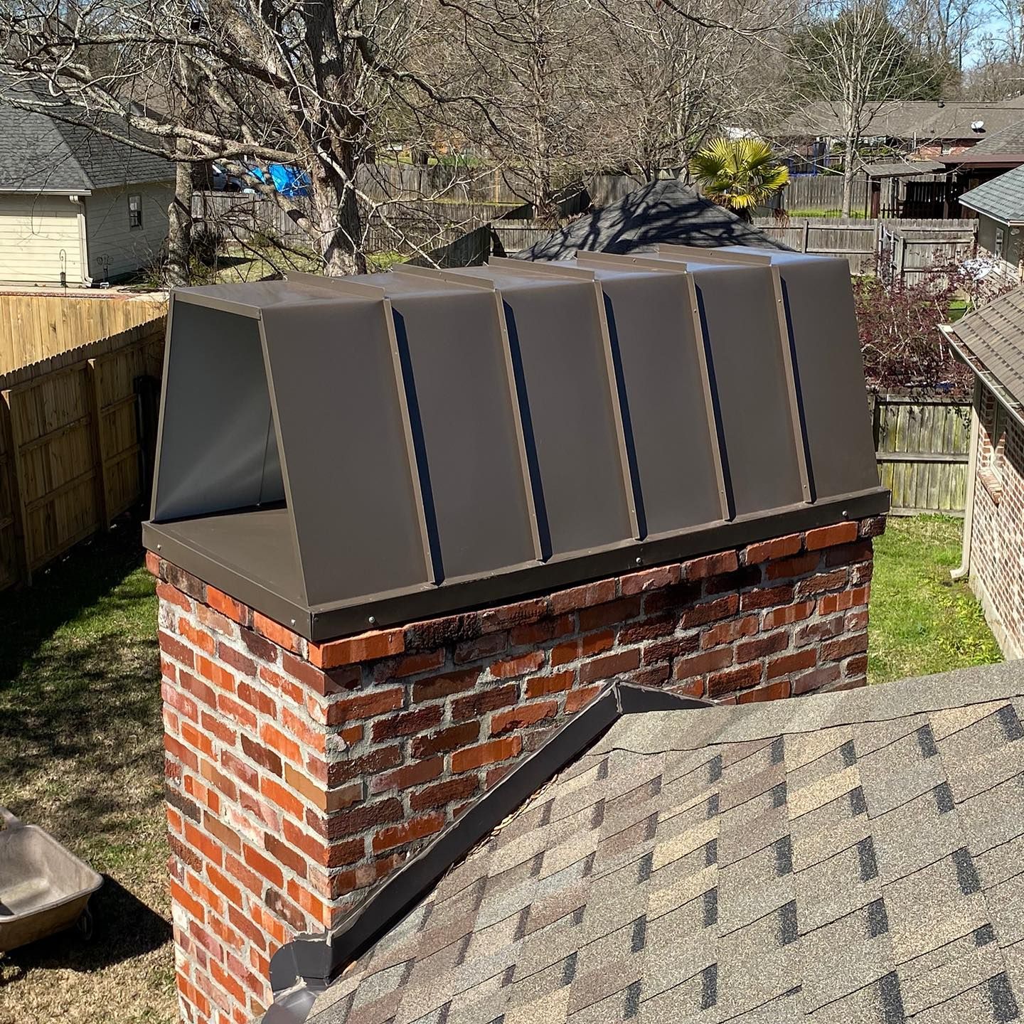 Brick chimney with brown metal cap and asphalt shingle roof, surrounded by houses and a wooden fence.