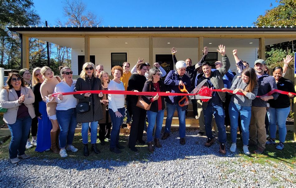 People cut a red ribbon in front of a white building, celebrating an event on a sunny day.