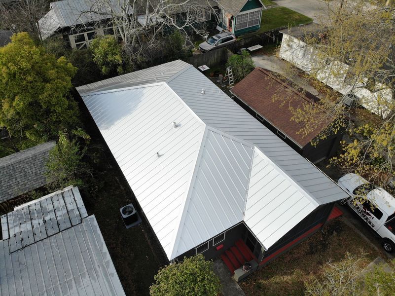 Aerial view of a house with a silver metal roof; surrounded by trees and other buildings.