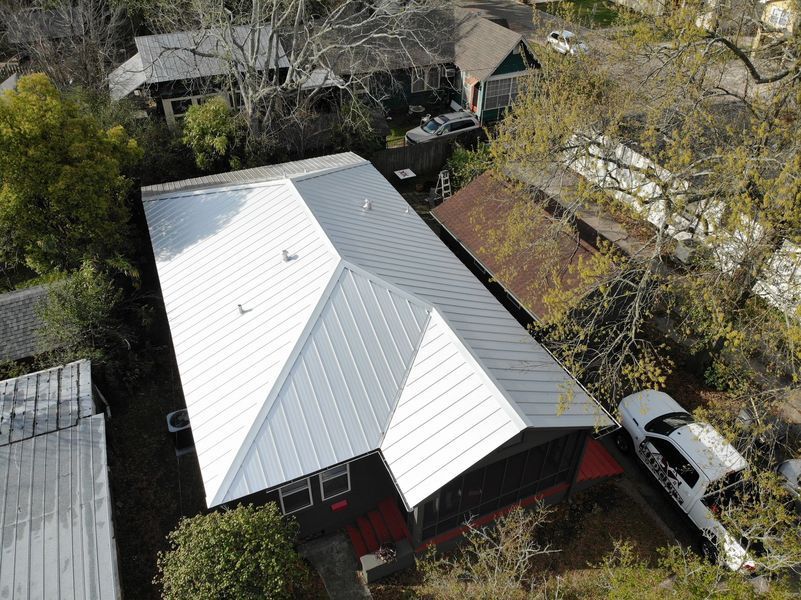Overhead view of a house with a silver metal roof, surrounded by trees and other buildings. A car is parked nearby.