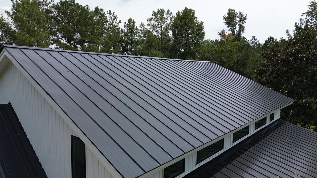 Black metal roof on a white building with windows, surrounded by green trees.