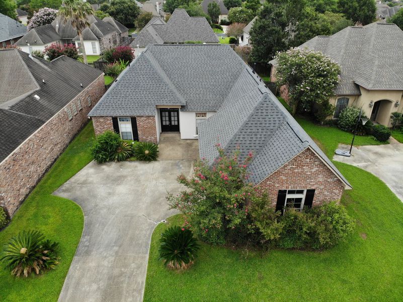 Drone shot of a brick house with a gray roof, surrounded by green lawn and other houses in a neighborhood.