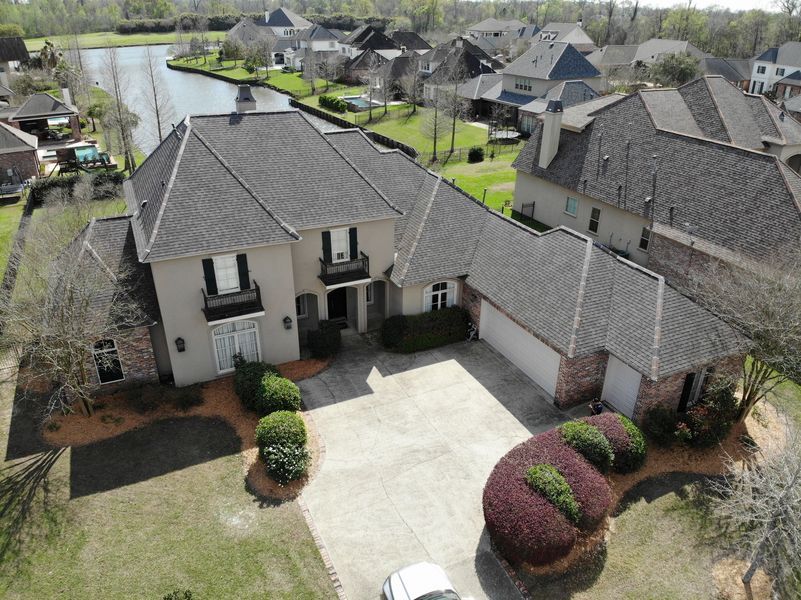 Drone view of a tan house with dark roof, two-car garage, and driveway, near a lake.