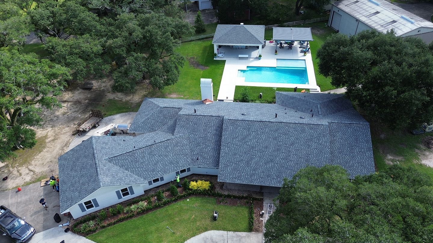 Aerial view of a gray house with a wavy roof, pool, and a white gazebo on green grass.