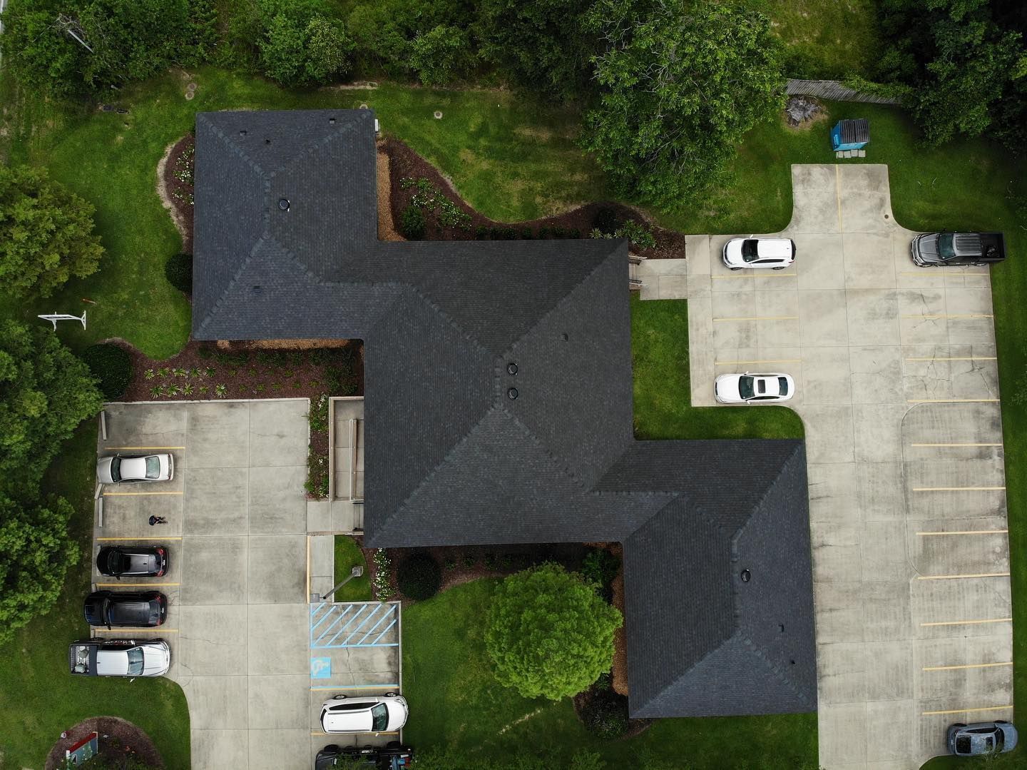 Aerial view of a building with a dark roof, surrounded by green lawn and parking lots with cars.