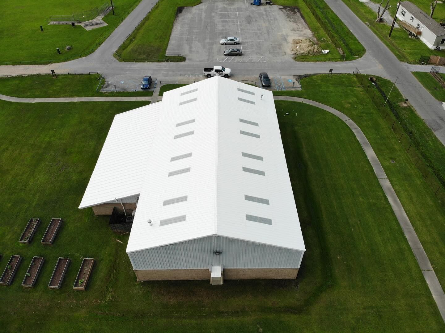 Aerial view: White-roofed building with rectangular vents on a grassy lot; adjacent parking lot and roads.