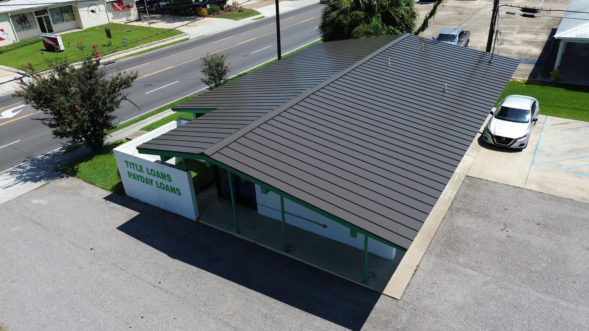Building with dark, curved roof, parking lot with white car. Road with cars visible.