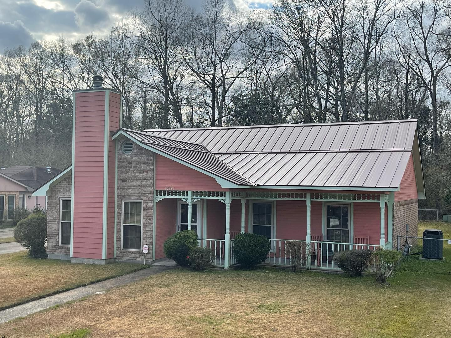 Pink house with metal roof, brick chimney, and small porch, set on a lawn with trees in the background.