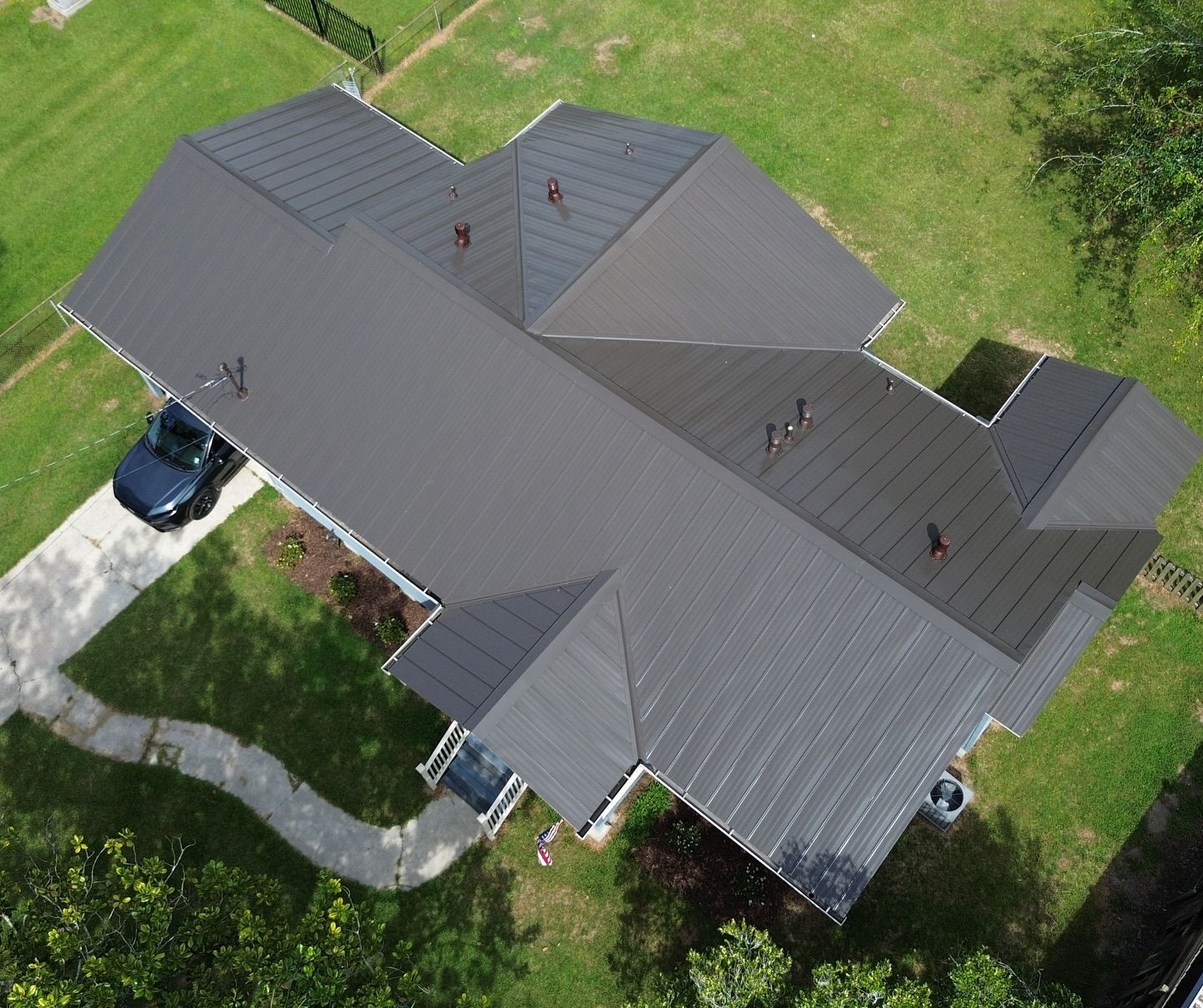 Aerial view of a house with a dark gray metal roof, surrounded by green grass and a driveway.