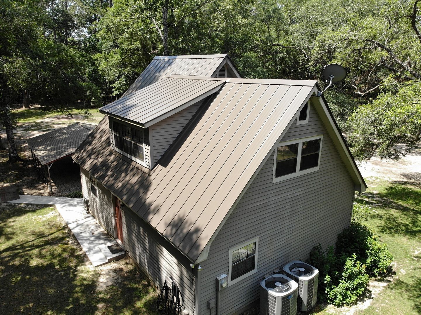 Tan-roofed house with two dormers, gray siding, set amid green trees. Two AC units below windows.