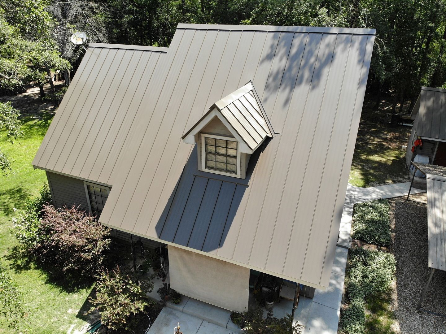 Beige house with dark gray metal roof, dormer window, surrounded by trees and greenery.