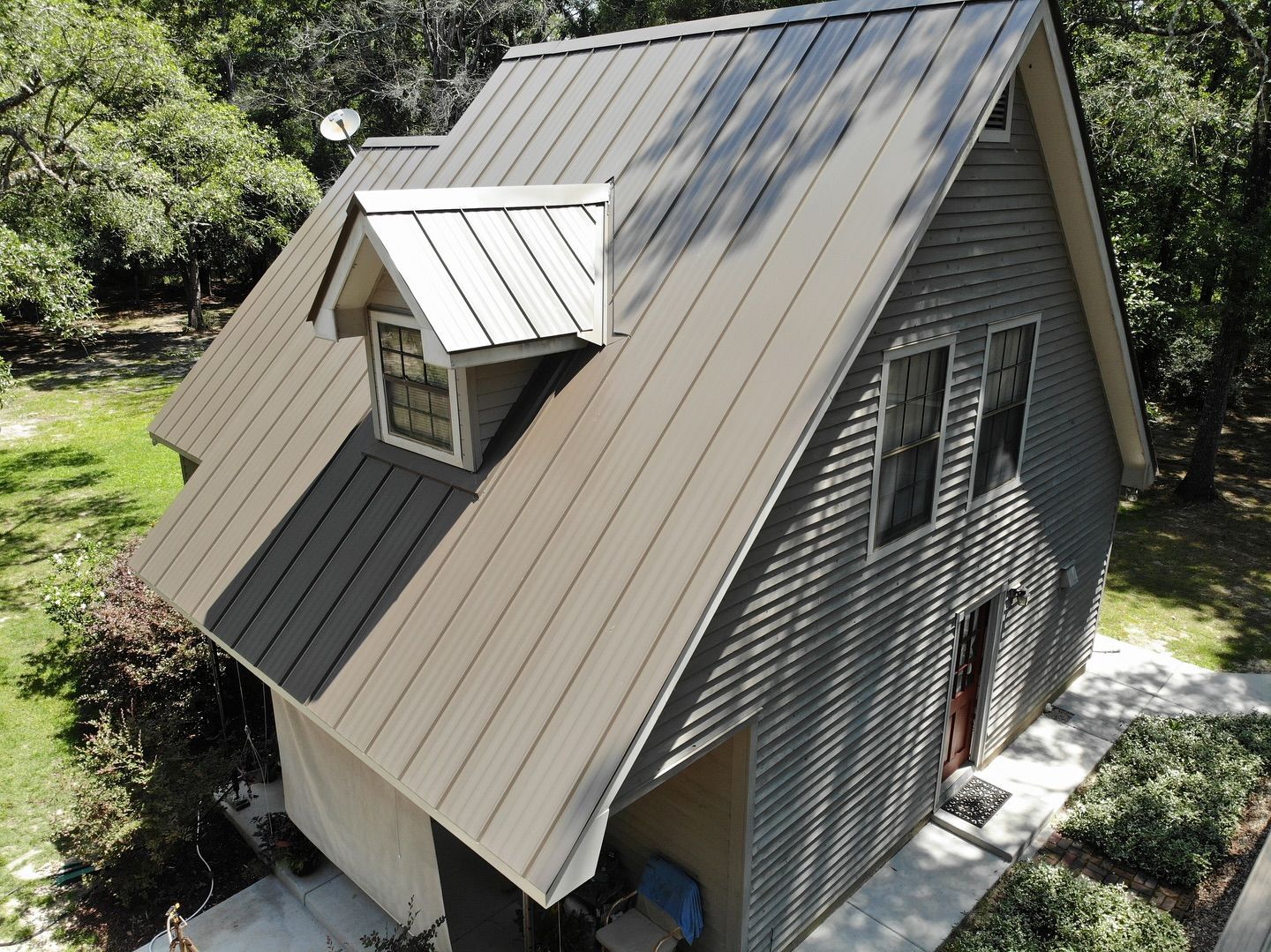A-frame building with tan metal roof, a dormer window, and gray siding, set among trees.