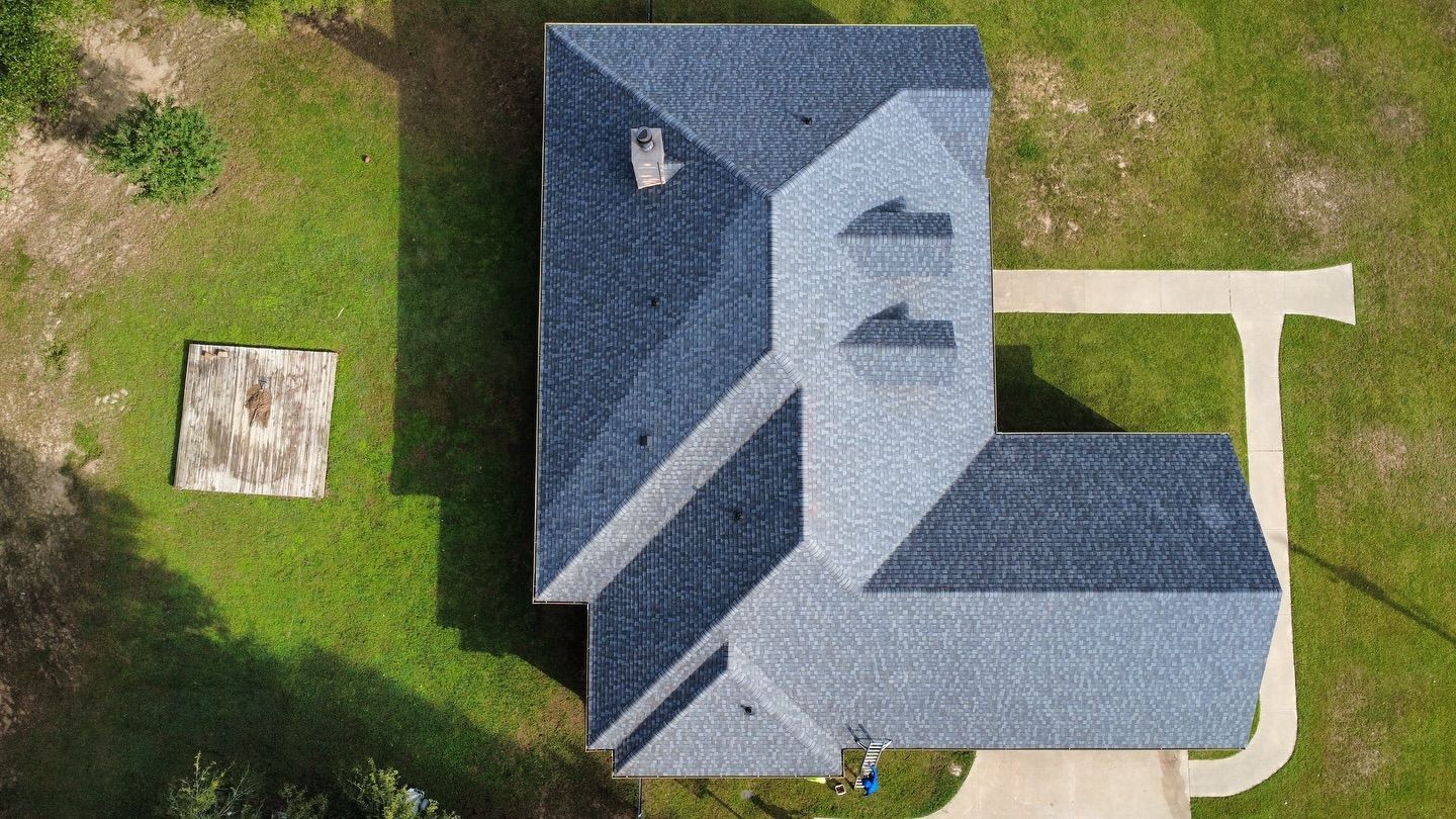 Overhead view of a house with a blue shingled roof, driveway, and grassy yard with a cement slab.