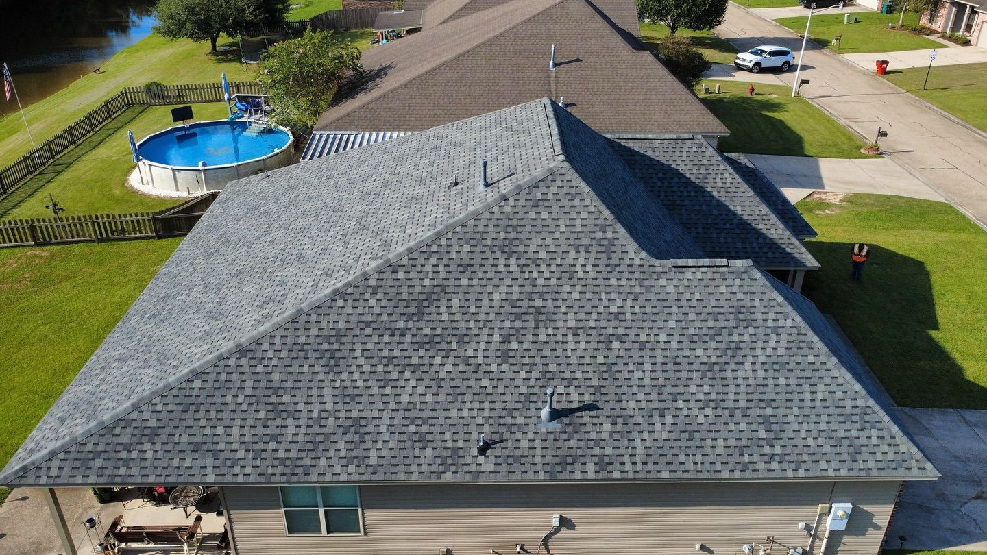 Aerial view of a house with a gray shingled roof, green lawn, and a pool, near a street with a white car.