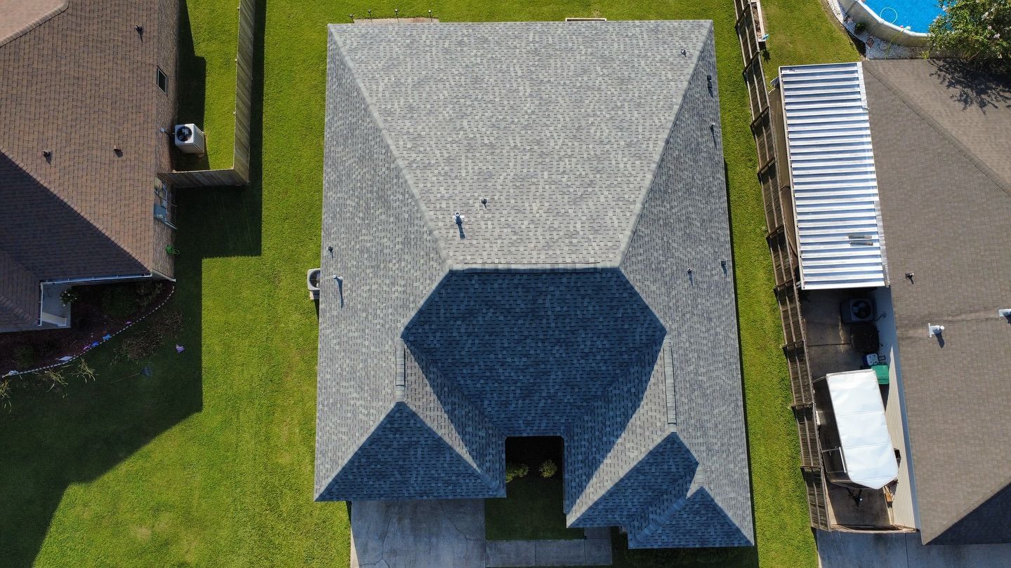 Overhead view of a gray roof with a complex shape, surrounded by green grass and neighboring houses.