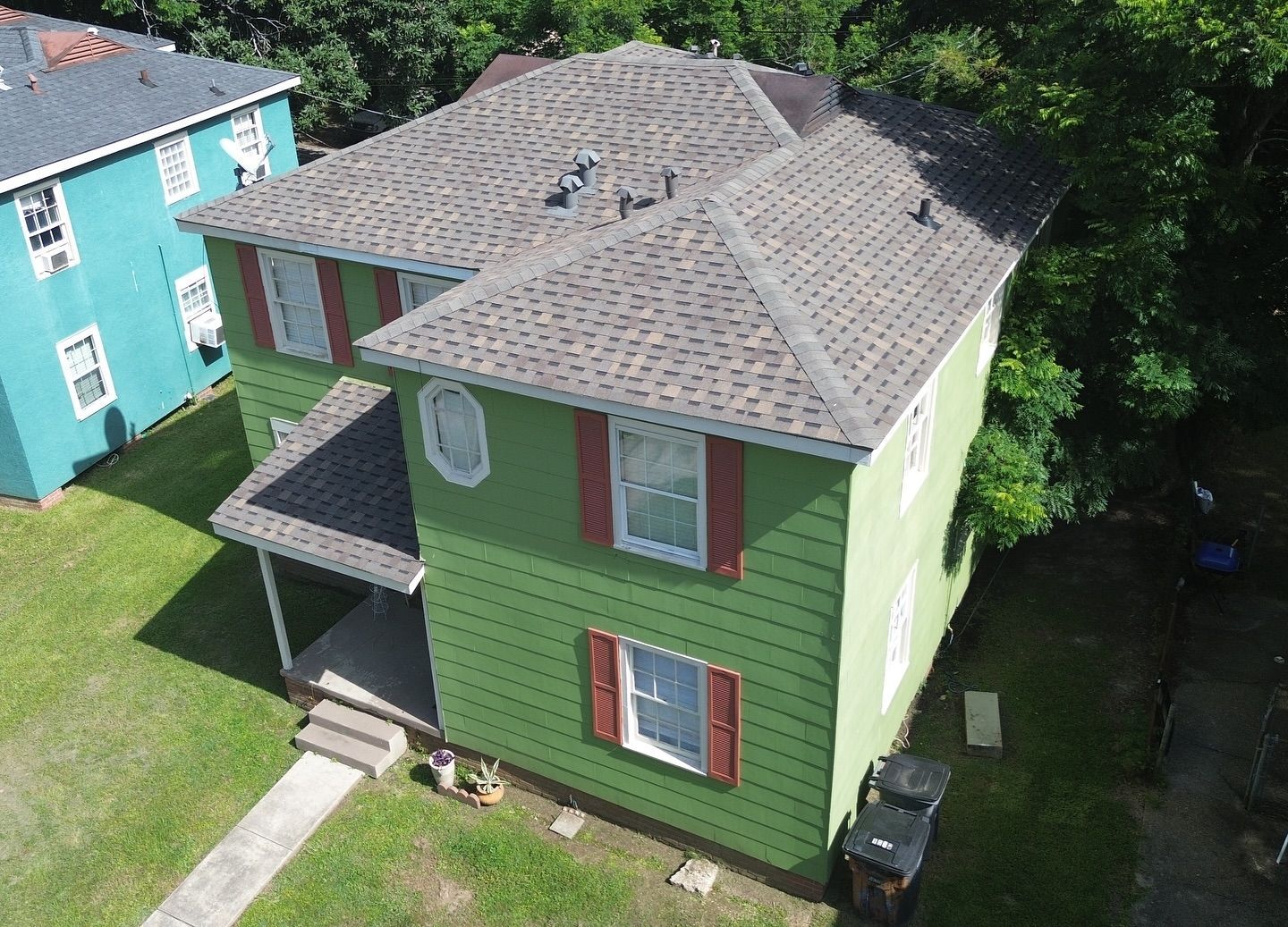 Green two-story house with brown roof and red shutters. Lawn and trees surround it, with a blue house to the left.