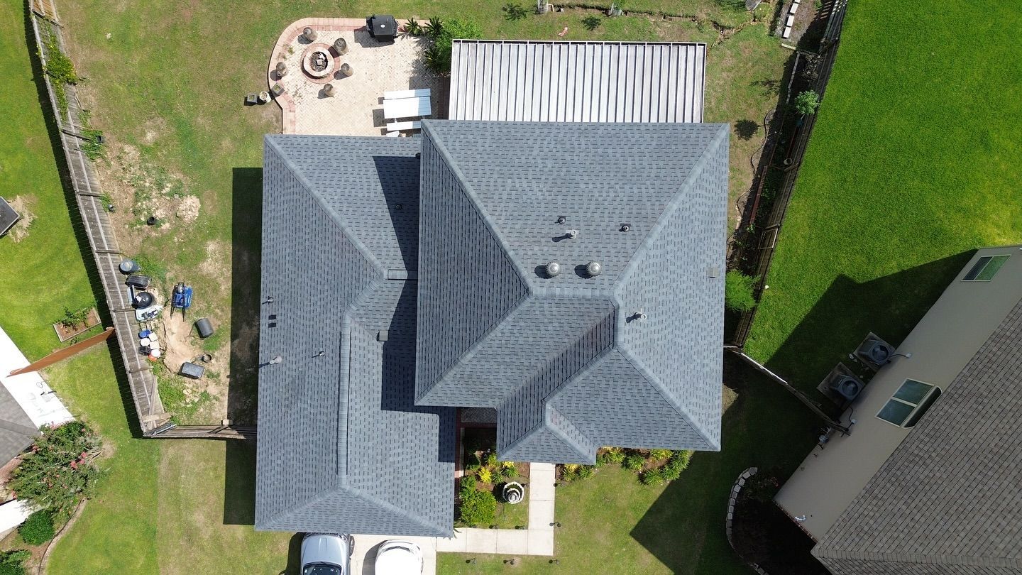 Overhead view of a house with a dark grey roof, surrounded by green grass and other houses in a sunny neighborhood.