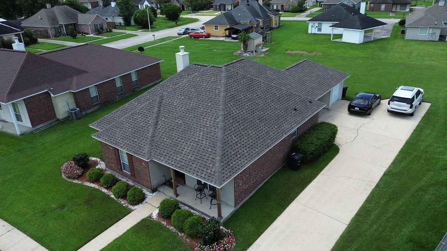 Aerial view of a brick house with a gray roof, driveway, and cars. Surrounded by green grass and other houses.