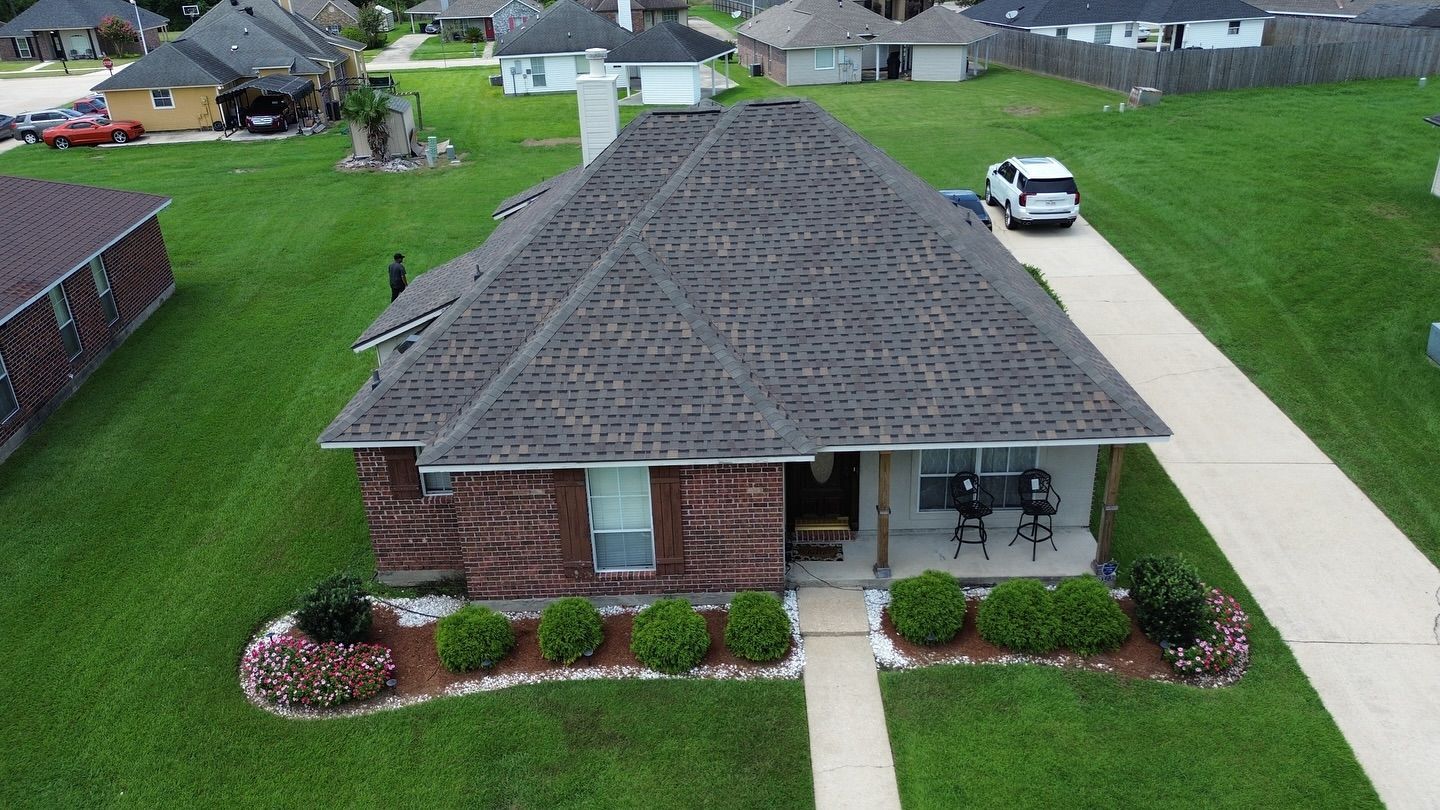 House with brown brick facade, dark roof, green lawn, and a driveway, with a white SUV parked.
