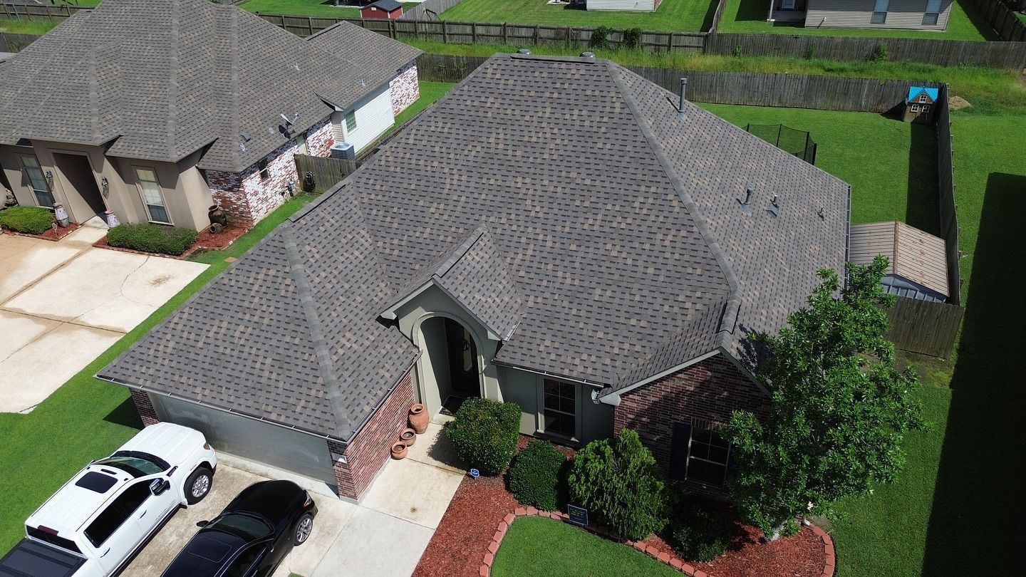 An aerial view of a house with a gray roof, brick facade, and a driveway with cars.