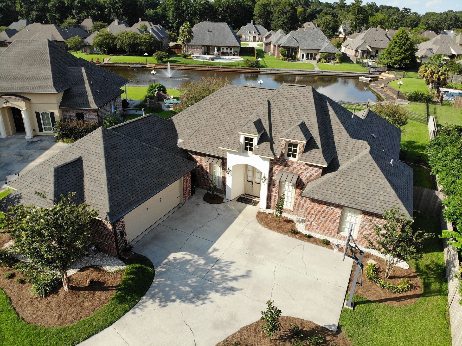 Brick house with dark roof, lake view, green grass, trees, and other houses.