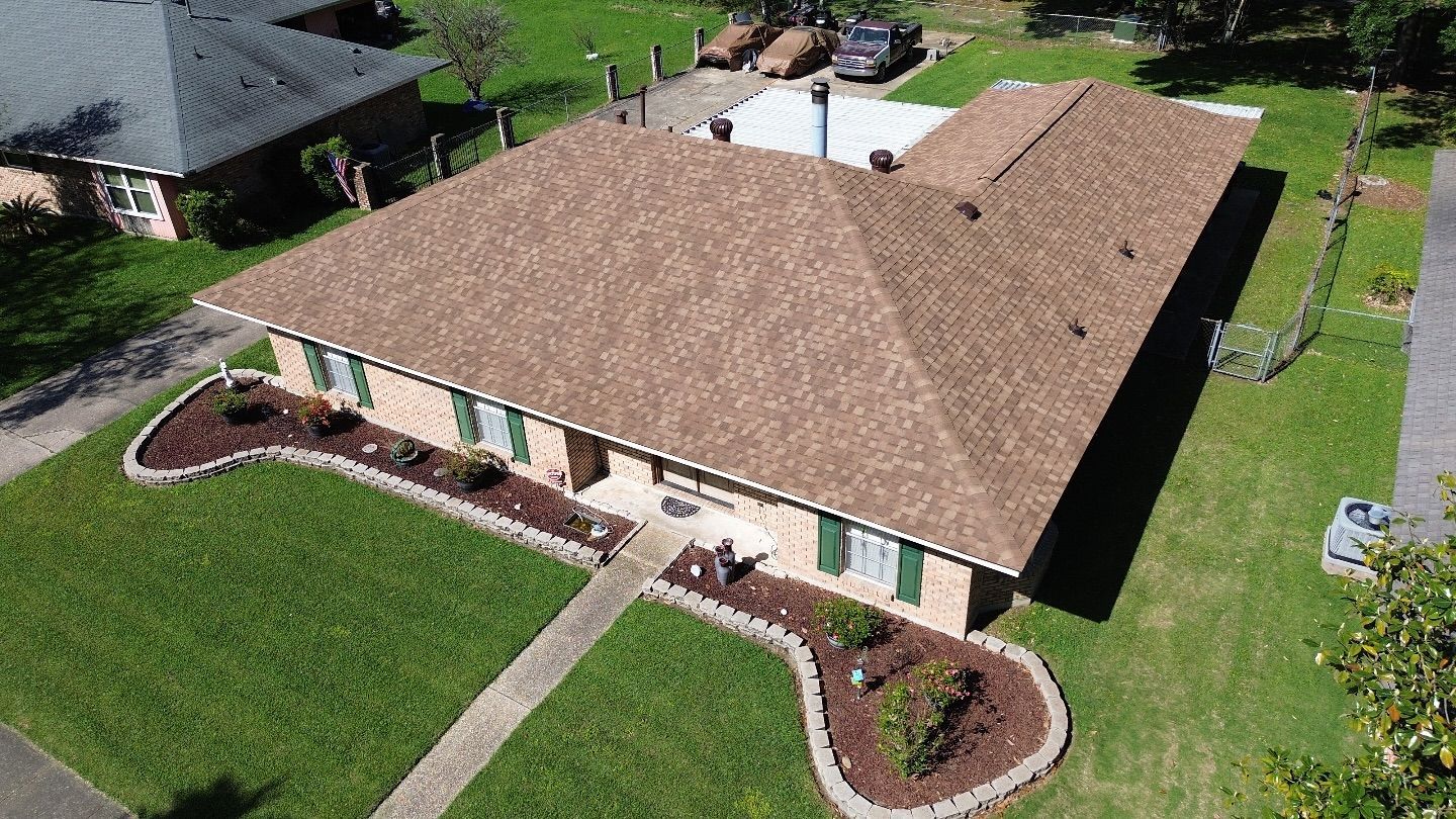 Brown-roofed house with green shutters and lawn. Landscaped front beds, surrounded by a walkway, in a suburban setting.