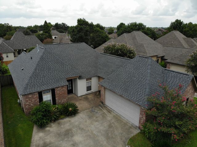 Drone view of a brick house with dark gray roof, garage, and a driveway, surrounded by trees and similar homes.