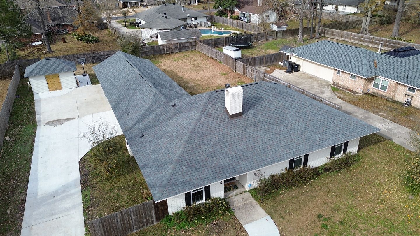 Aerial view of a white house with a dark gray roof and a long driveway in a suburban neighborhood.