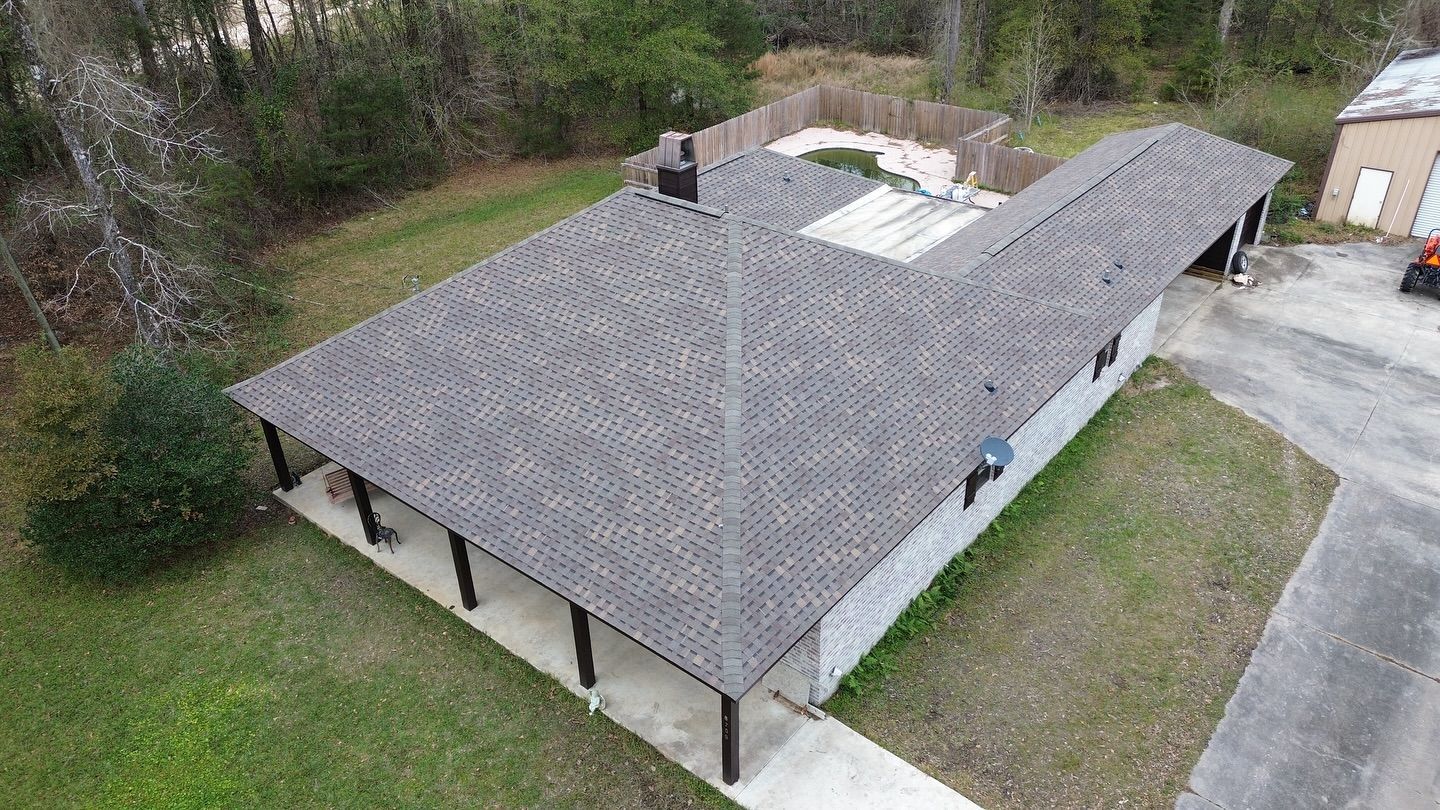 Aerial view of a single-story building with a dark gray roof and porch. Surrounded by grass and trees.
