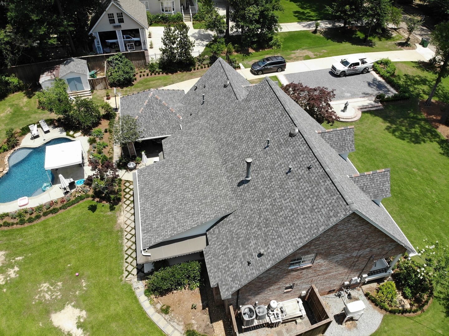 Aerial view of a house with a gray roof, a pool, and a car parked on a driveway surrounded by green grass.