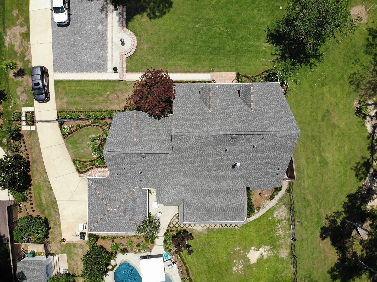 Aerial view of a house with a grey roof, driveway, cars, and surrounding green lawn and trees.