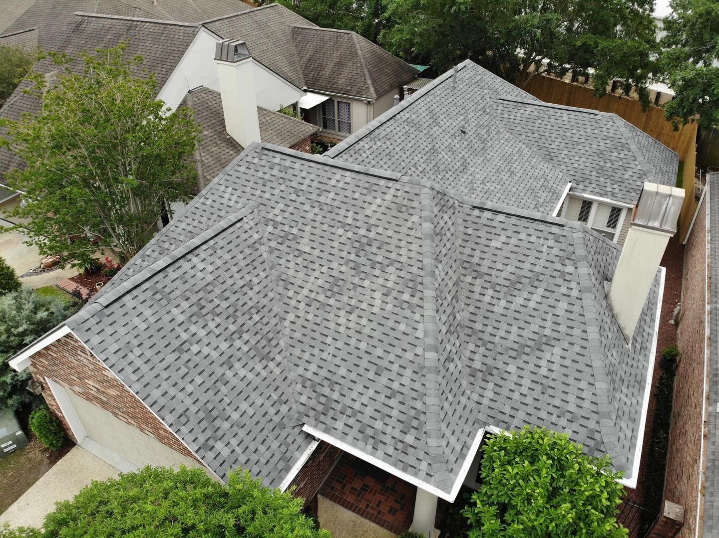 Overhead view of a gray shingled roof with a chimney, bordered by trees and a brick facade.