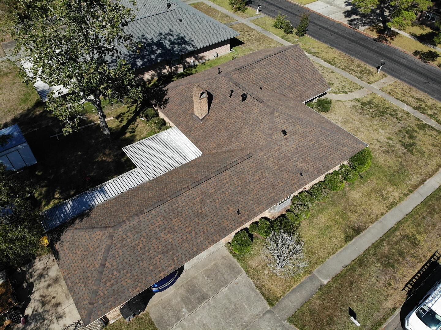 Aerial view of a house with a brown roof, chimney, and driveway, surrounded by grass and a tree.