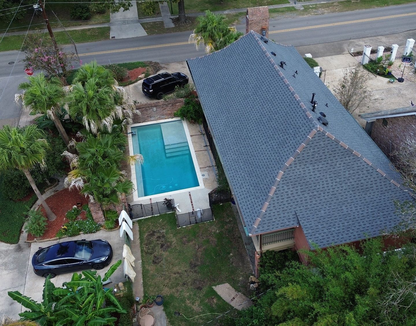 Aerial view of a house with a pool, two cars, and a gray roof, surrounded by trees and a fence.