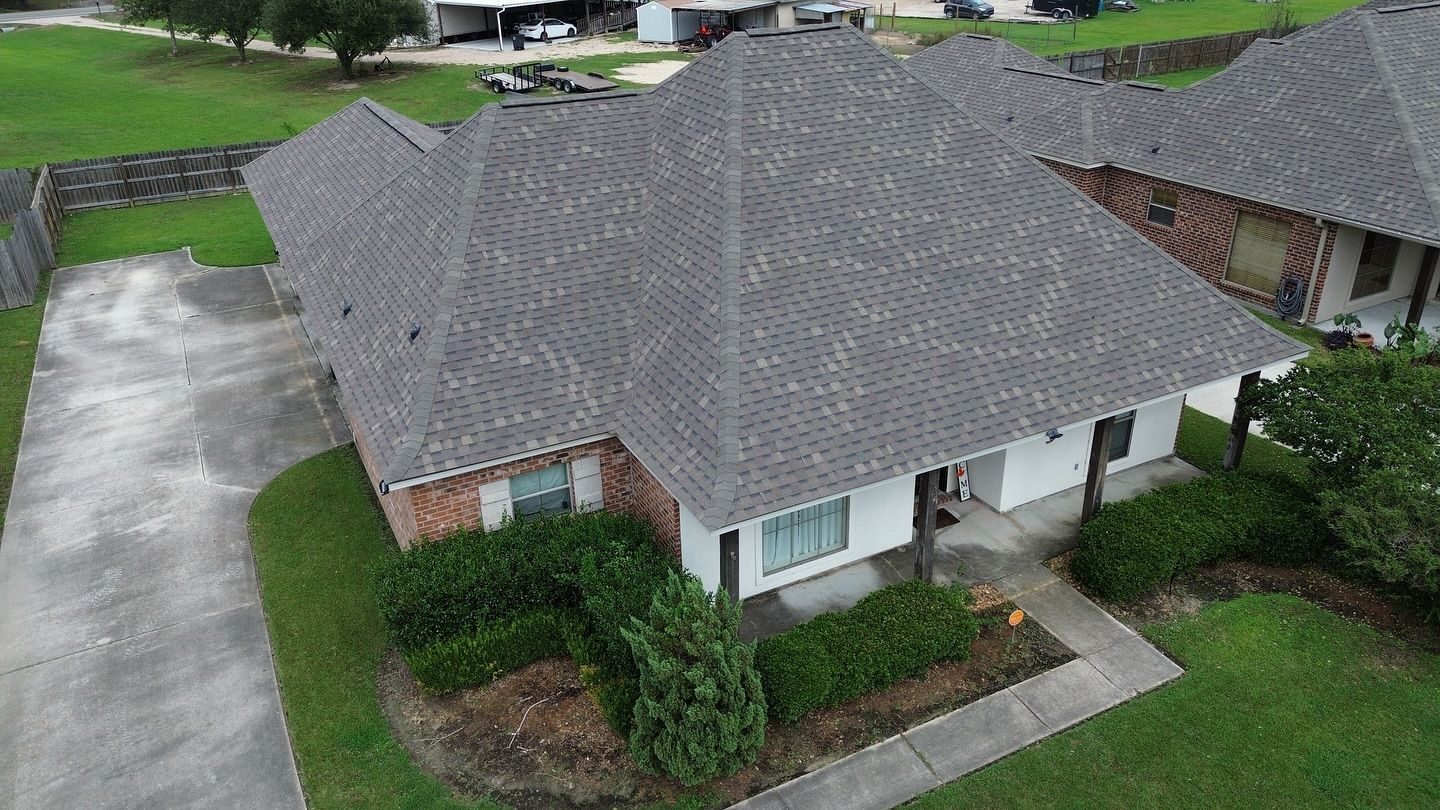 A one-story house with gray shingles and a white facade, surrounded by green grass and a driveway.