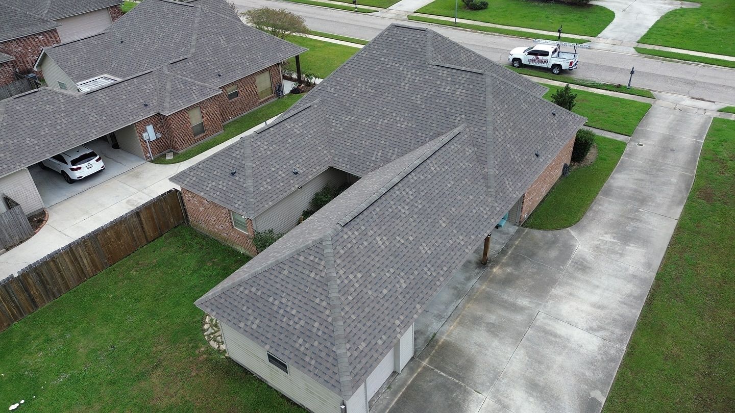 Overhead view of a gray-roofed house, driveway, green lawn, and neighboring houses in a suburban setting.
