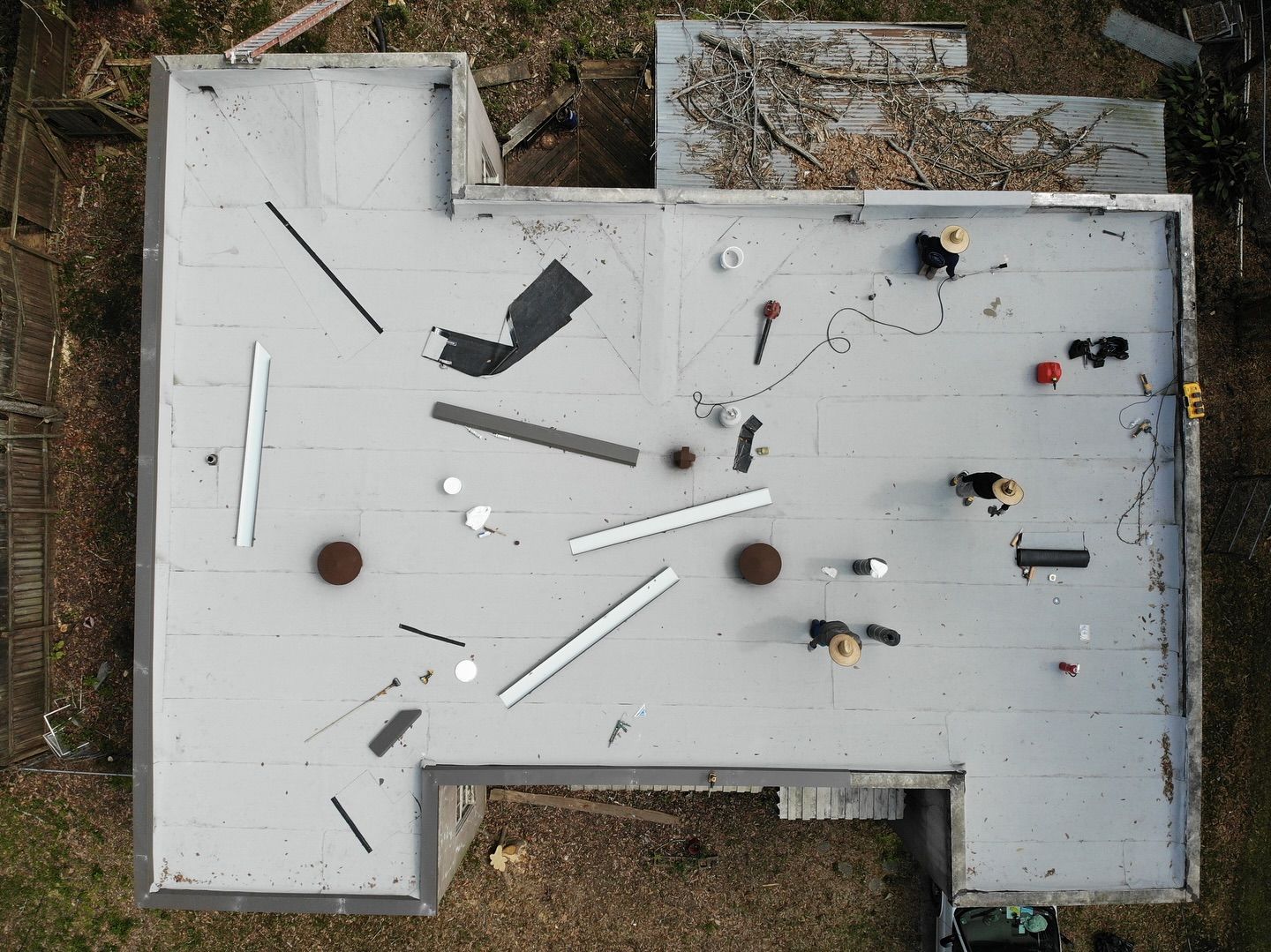Overhead view of workers on a gray flat roof with tools, debris, and two brown metal vents.