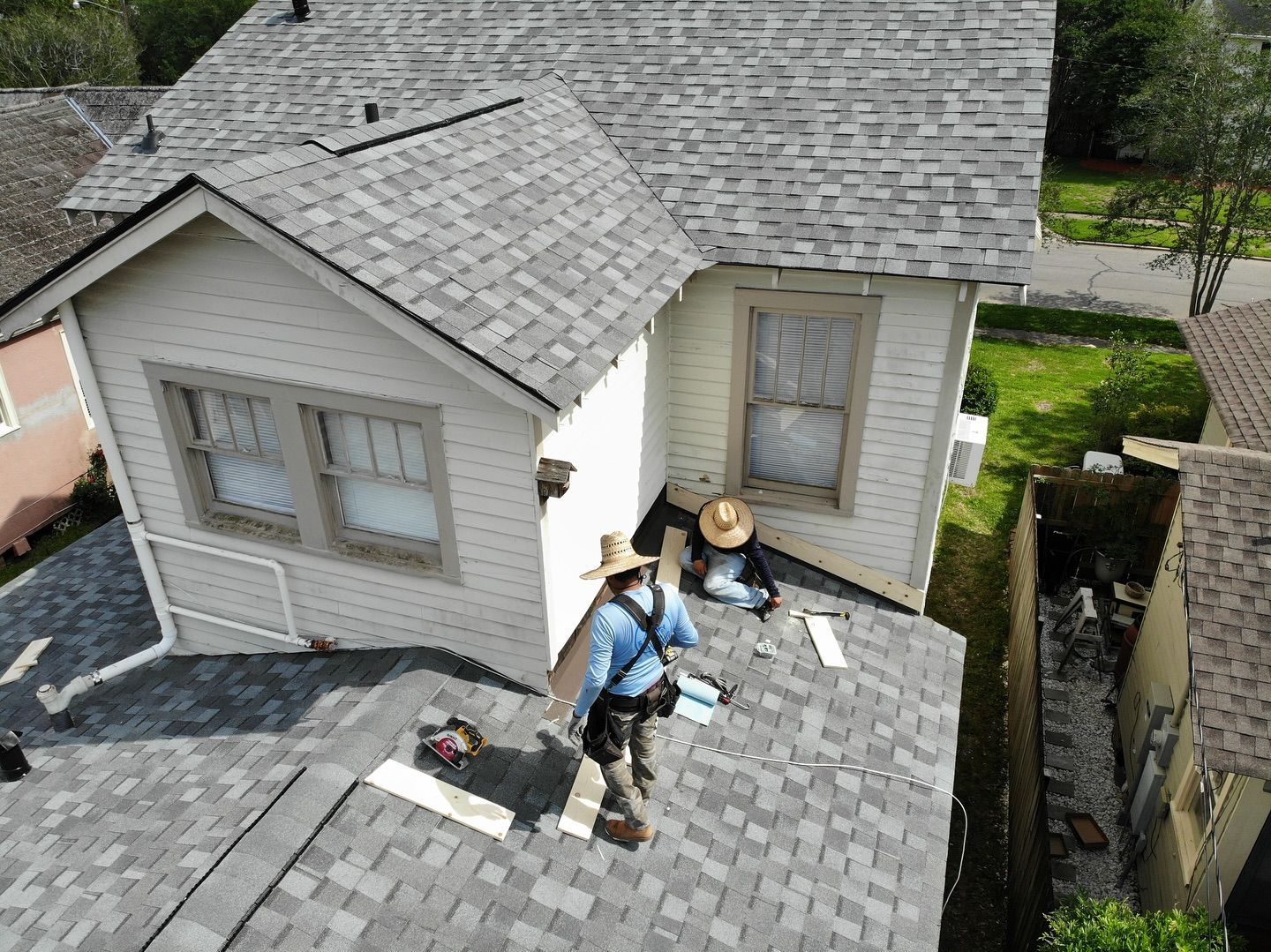 Roofers working on a gray-shingled roof of a white house. They are wearing hats and tool belts.