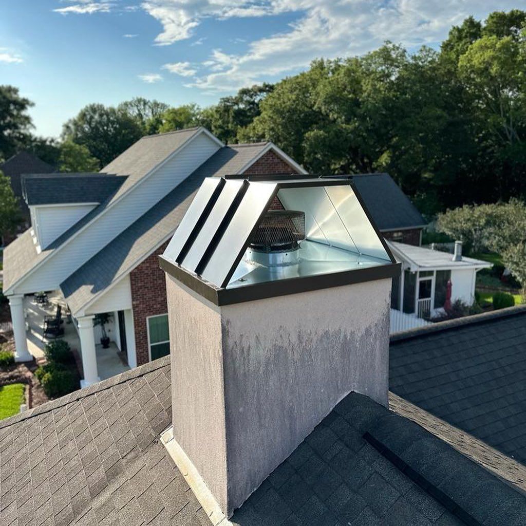 Chimney on a house roof with a metallic cap, surrounded by trees, under a blue sky.