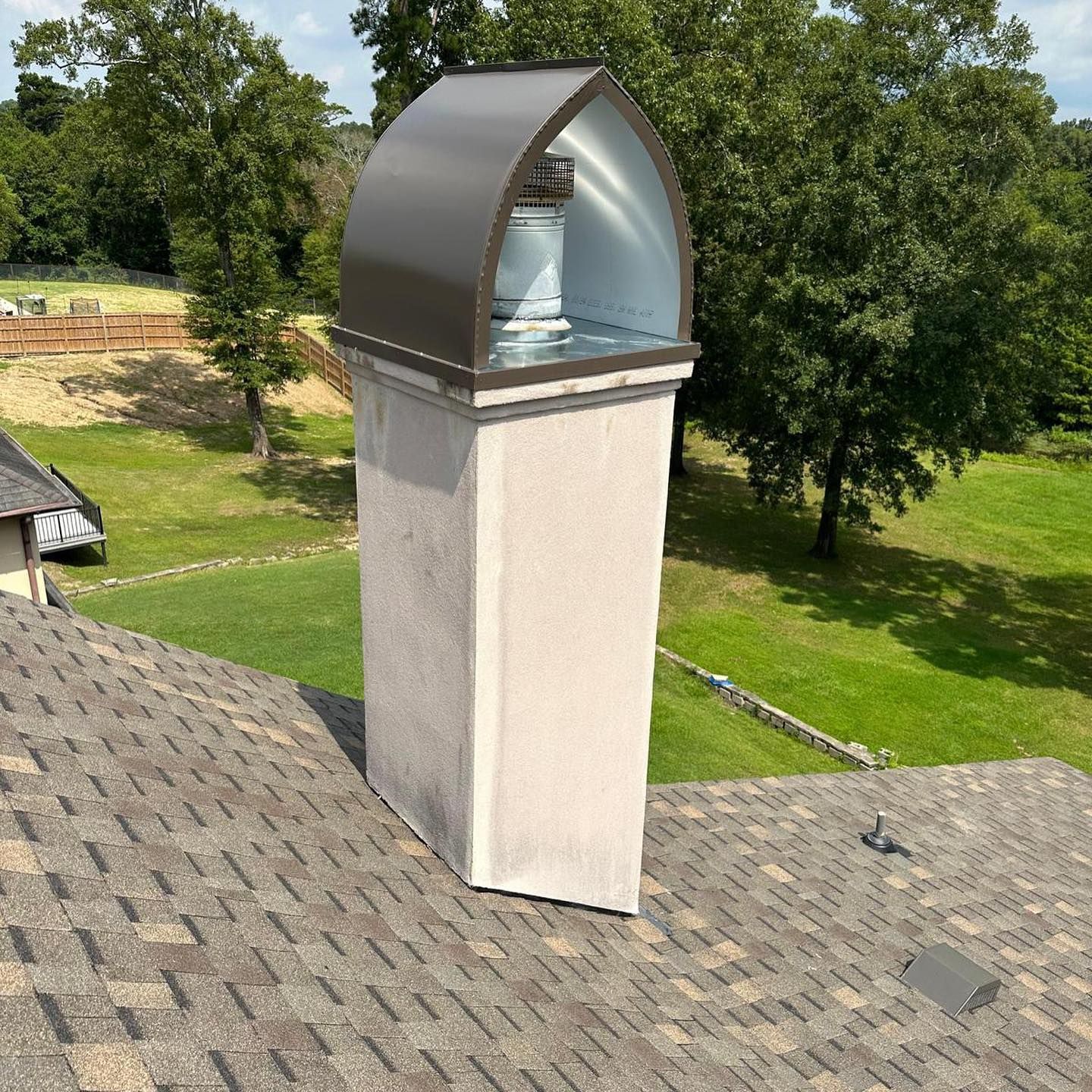 A chimney on a roof, with a metal arched cap; trees and grass in the background.