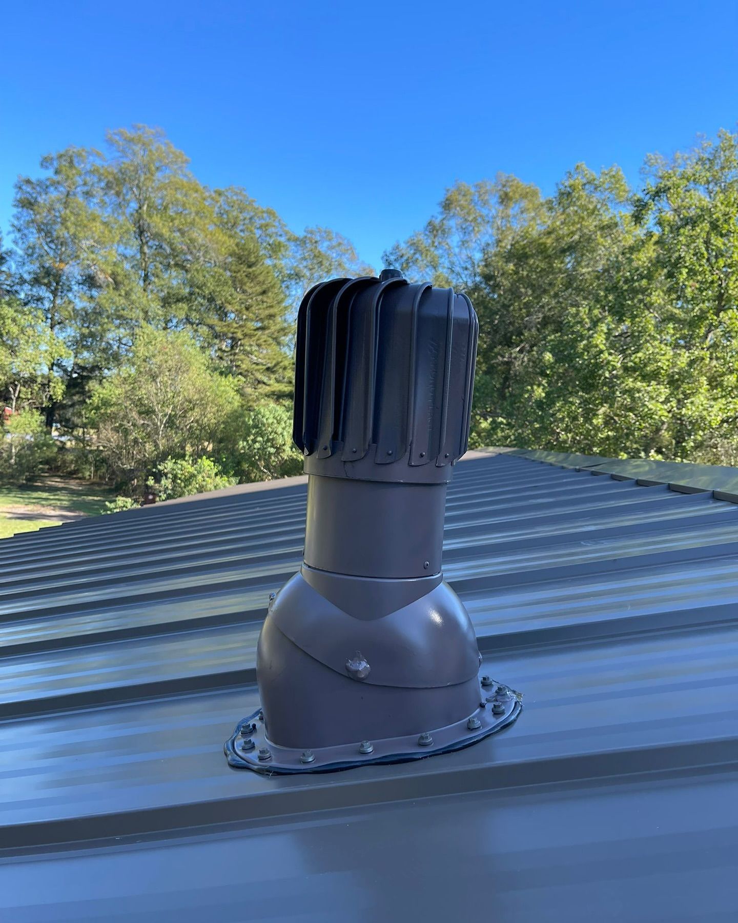 Gray roof vent on a metal roof with trees and blue sky in the background.