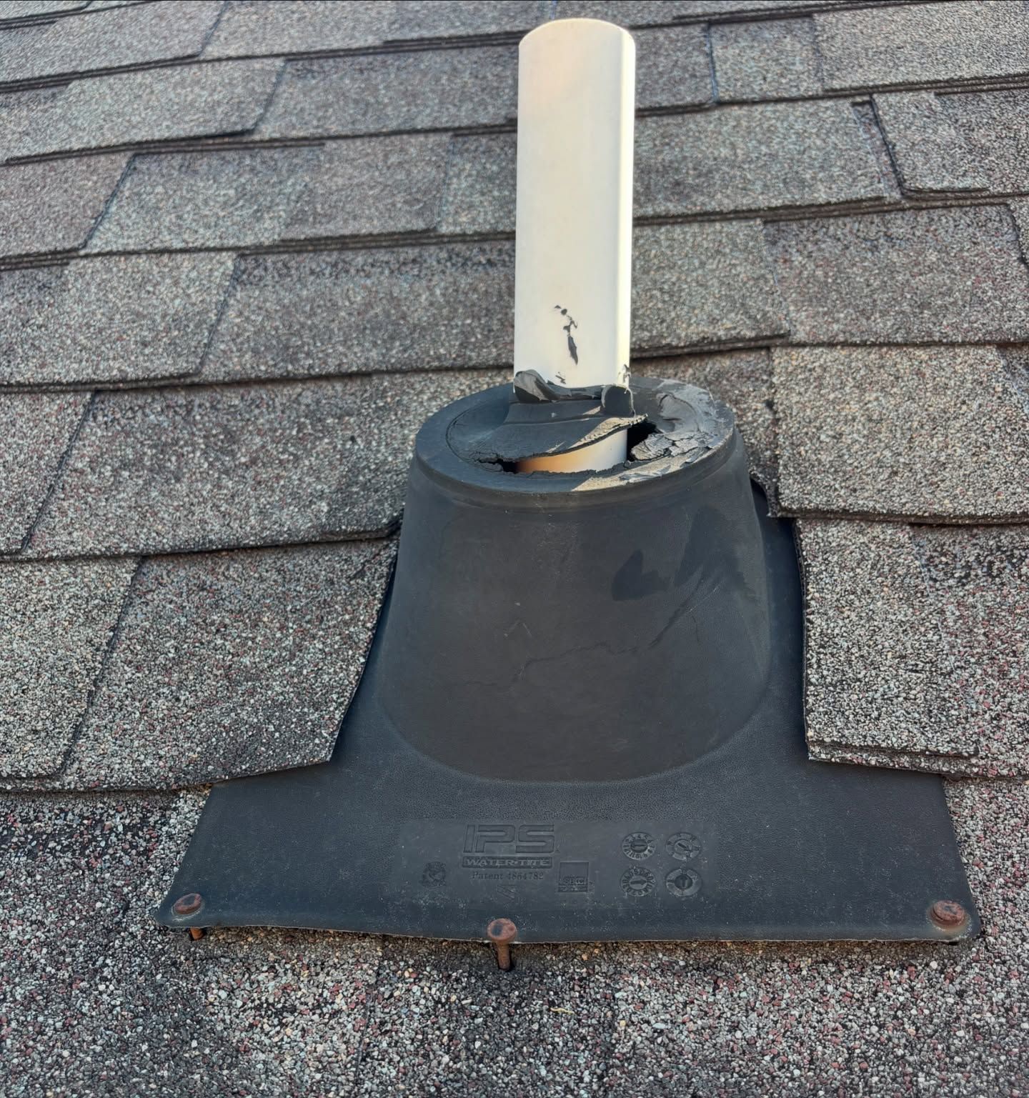 Damaged black roof vent with a white pipe extending upward, surrounded by asphalt shingles.