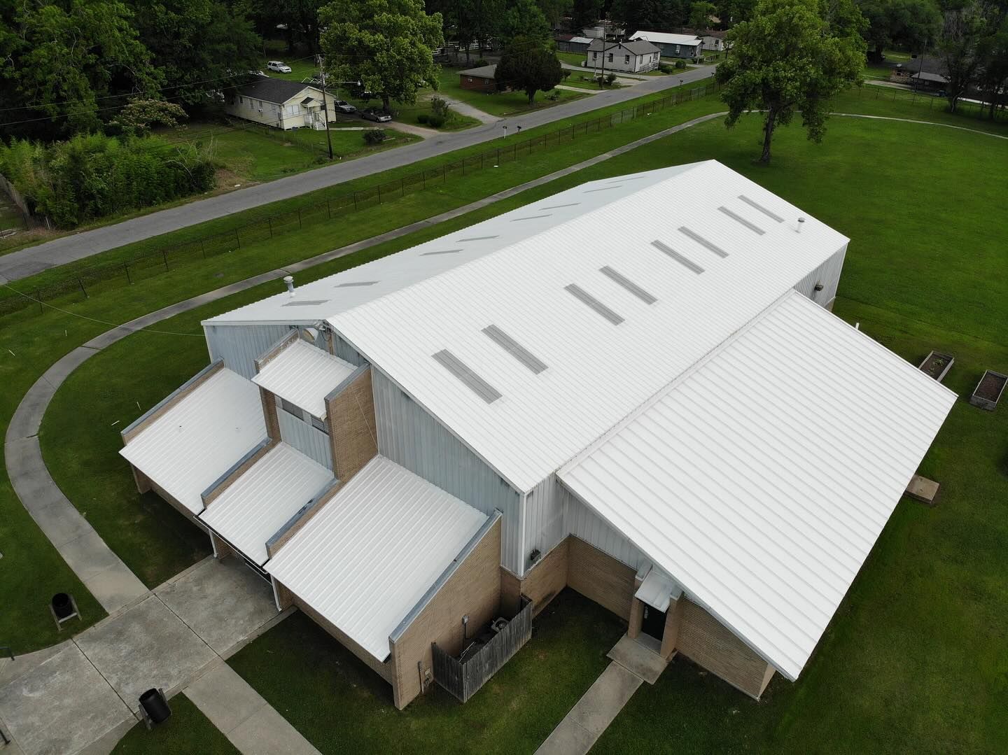 Aerial view of a white-roofed building with rectangular skylights; brown brick walls, green grass, and a pathway.