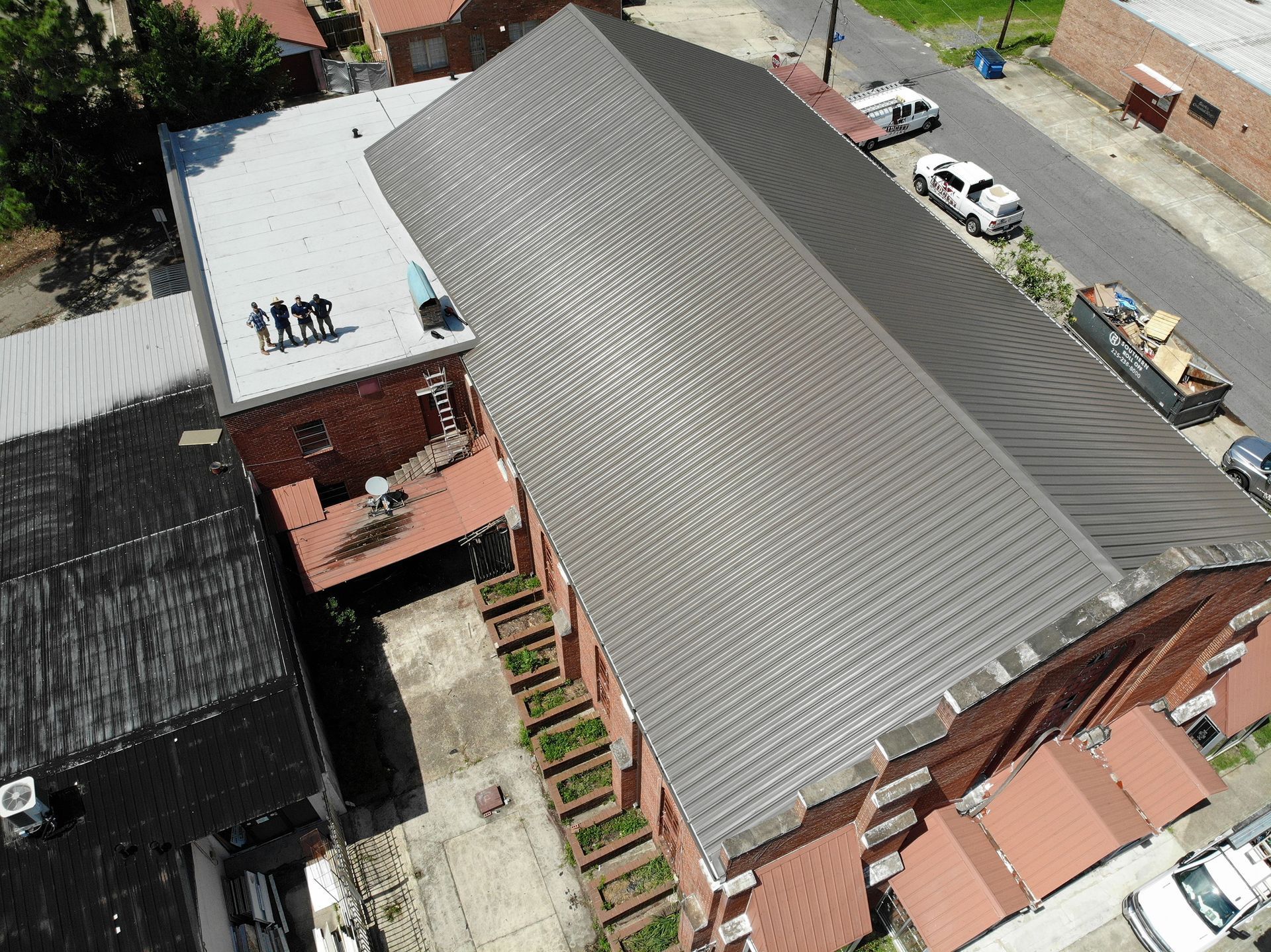 Aerial view of a brick building with a gray roof and a flat roof. Several people stand on the flat roof.