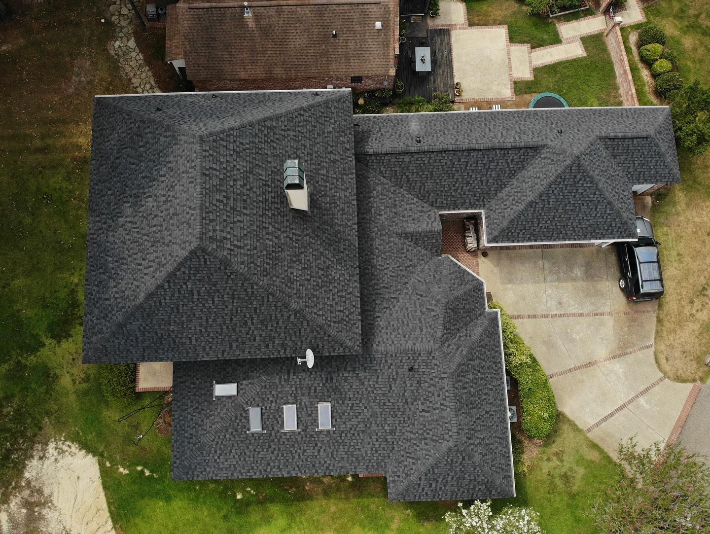 Aerial view of a dark gray asphalt shingled roof on a house, with driveway and nearby lawns.
