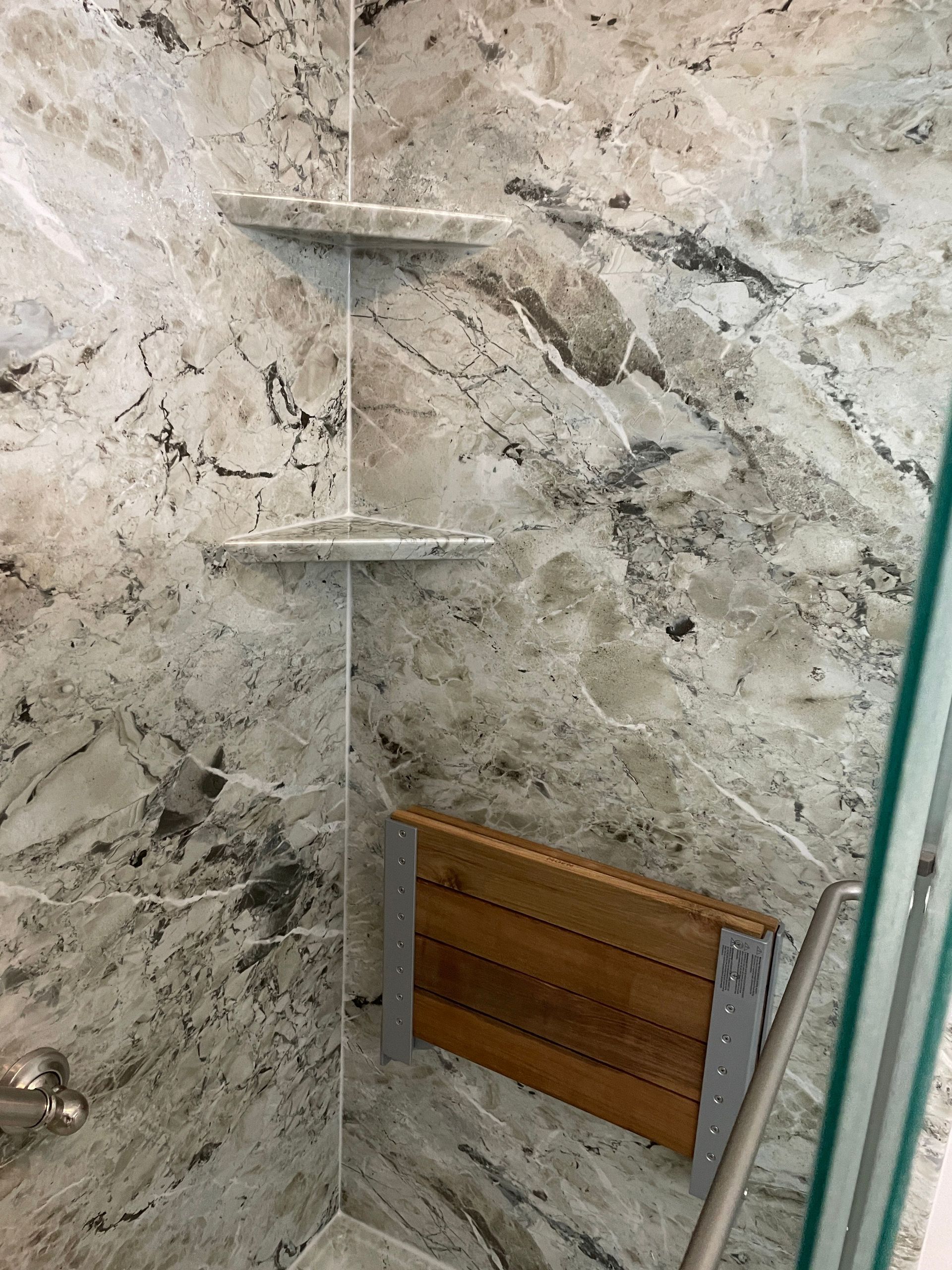 Corner shower with marble-look tile, glass door edge, corner shelves, and a wooden bath accessory holder