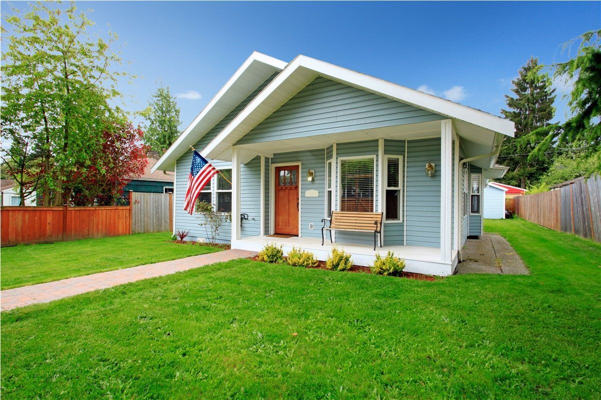 Small white house with a porch, American flag, and bright green lawn under a blue sky