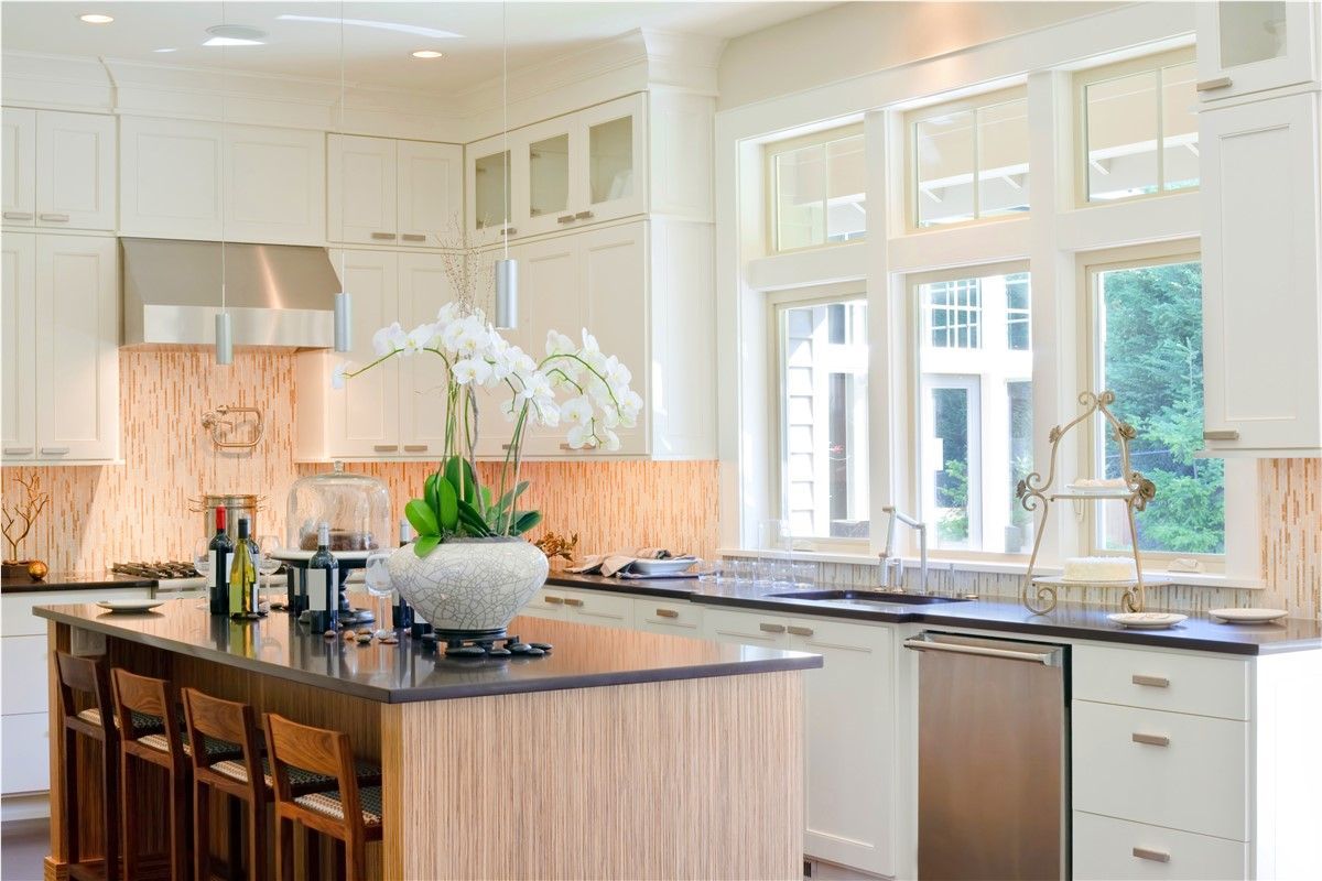 Bright, modern kitchen with a large island, wooden stools, and white cabinets near tall windows.