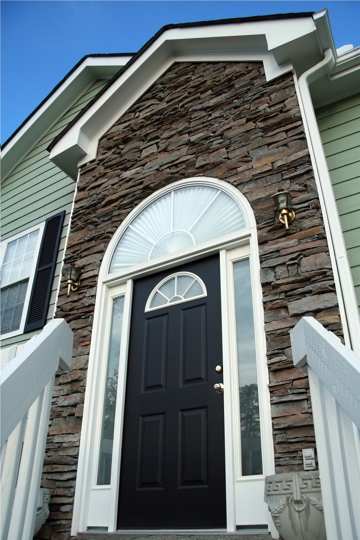 Front entrance with black door, white trim, and stone facade on a house under a blue sky
