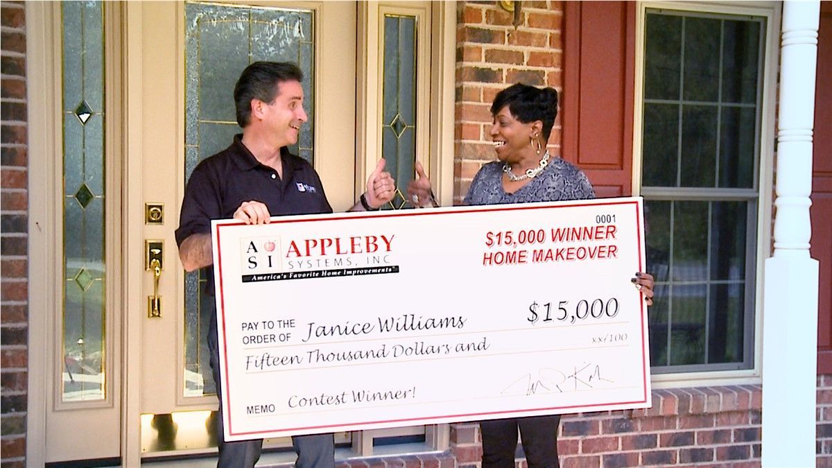 Two men hold an oversized check outside a brick house doorway.